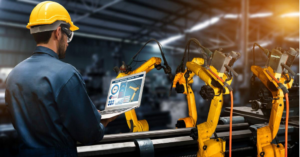 Man with construction hat on holding laptop computer in front of robotic machinery.