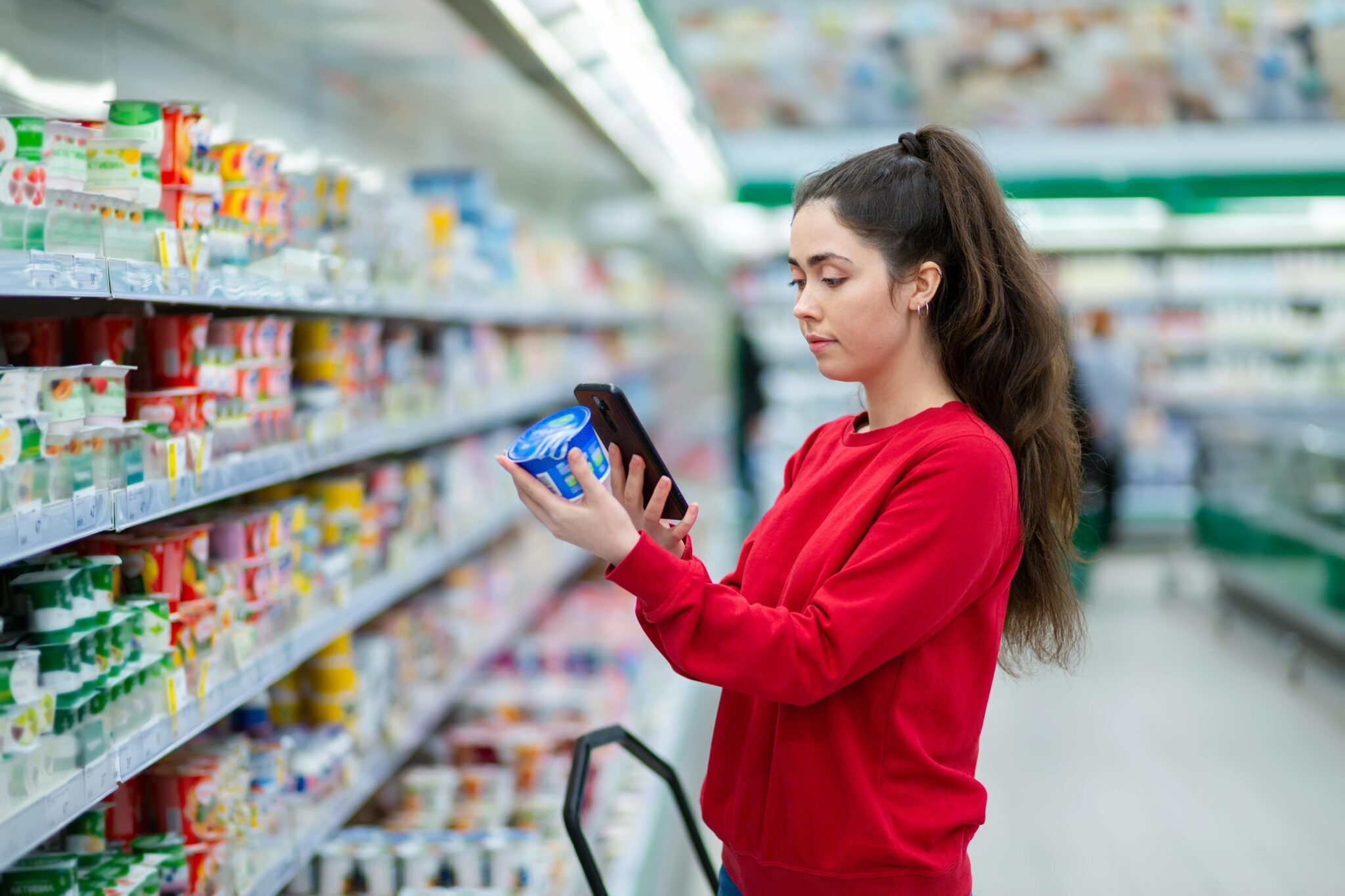 Woman in grocery store holding a product in one hand and taking a picture of the product with her mobile phone in the other