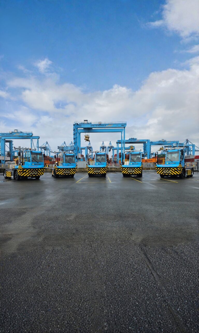Five blue terminal tractors lined up at a shipping port, with container cranes and cargo containers visible in the background under a partly cloudy sky.