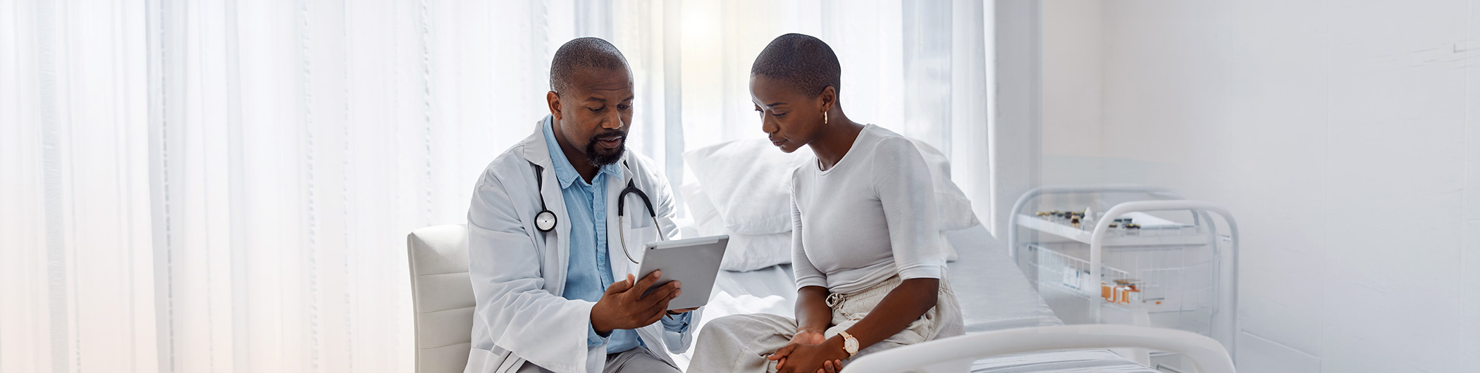 Doctor showing information on a tablet to a patient seated on a hospital bed in a bright clinical room.